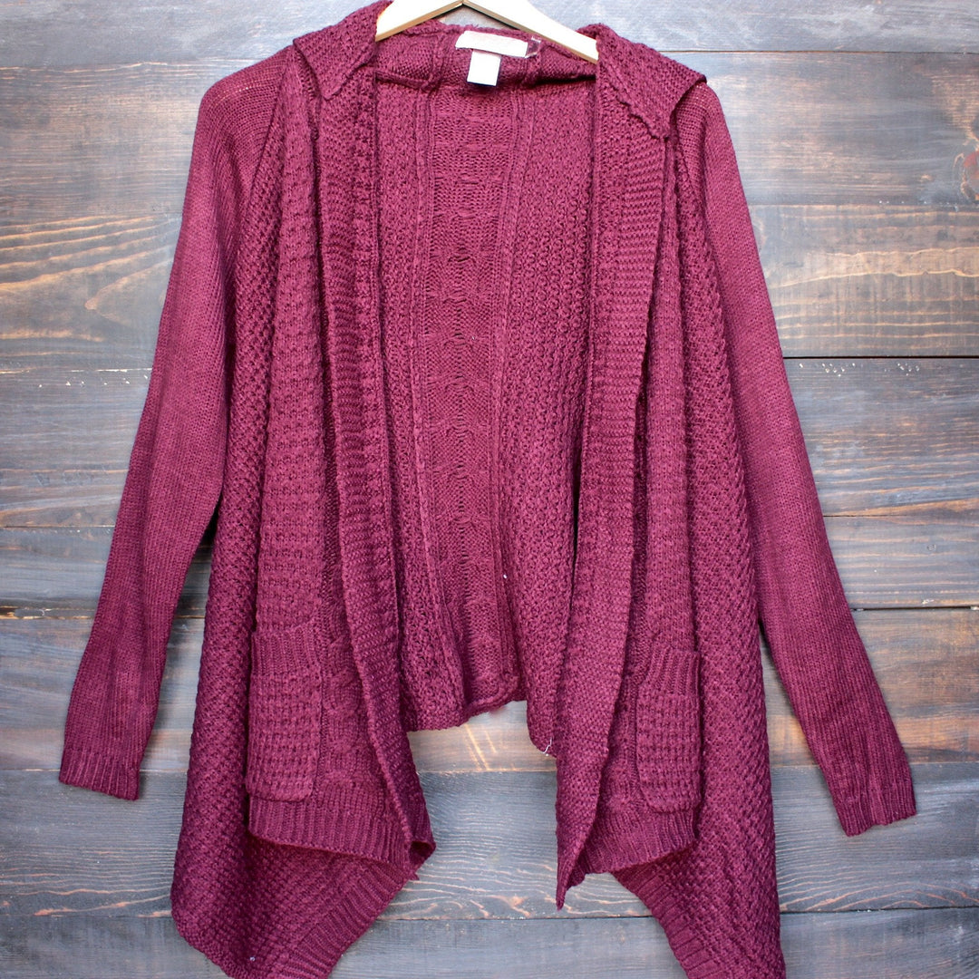 Burgundy open-front knit cardigan with hood and two front pockets, hanging against a wood backdrop