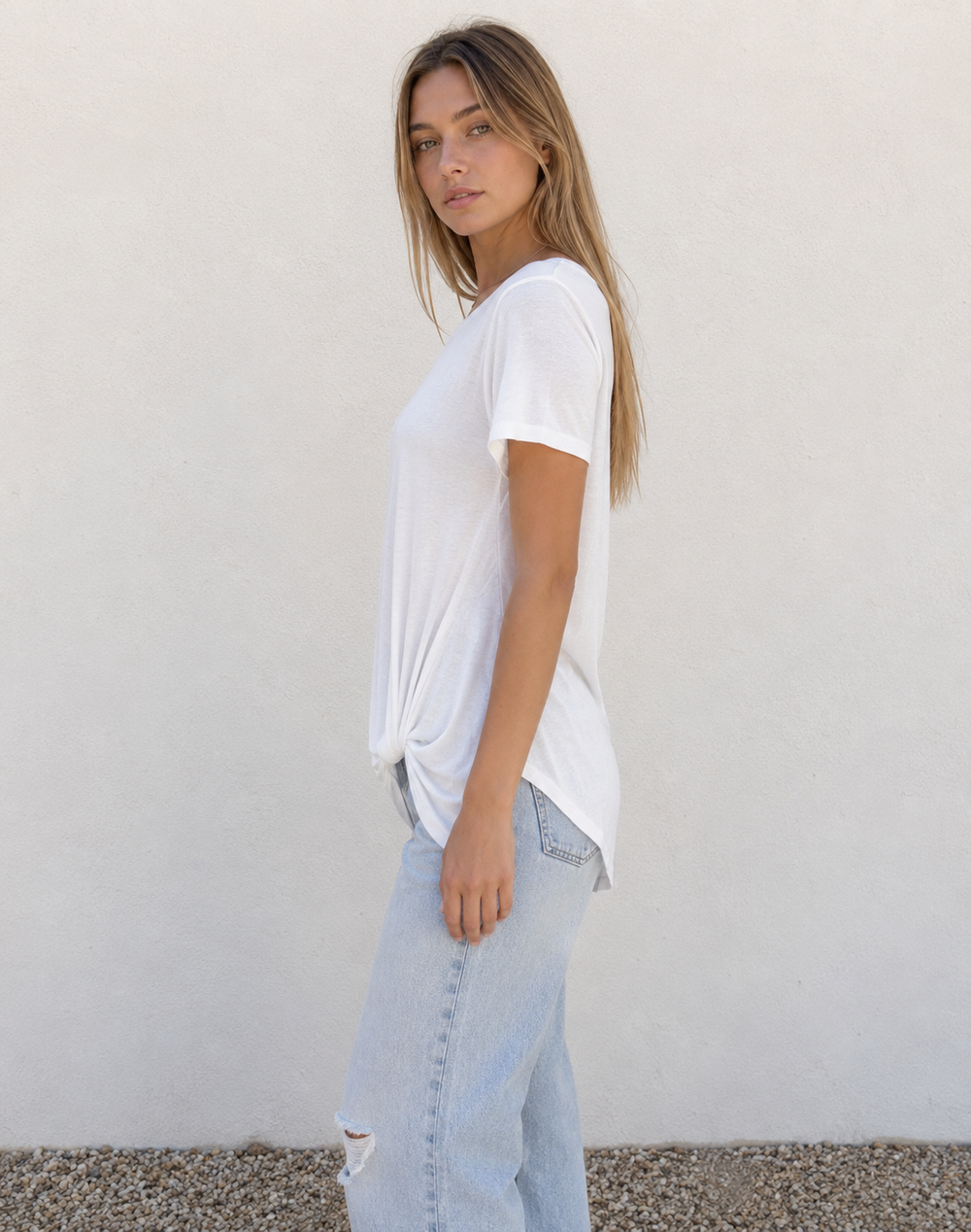 A woman wearing a white short-sleeve t-shirt and light-wash distressed jeans, standing against a plain off-white wall.