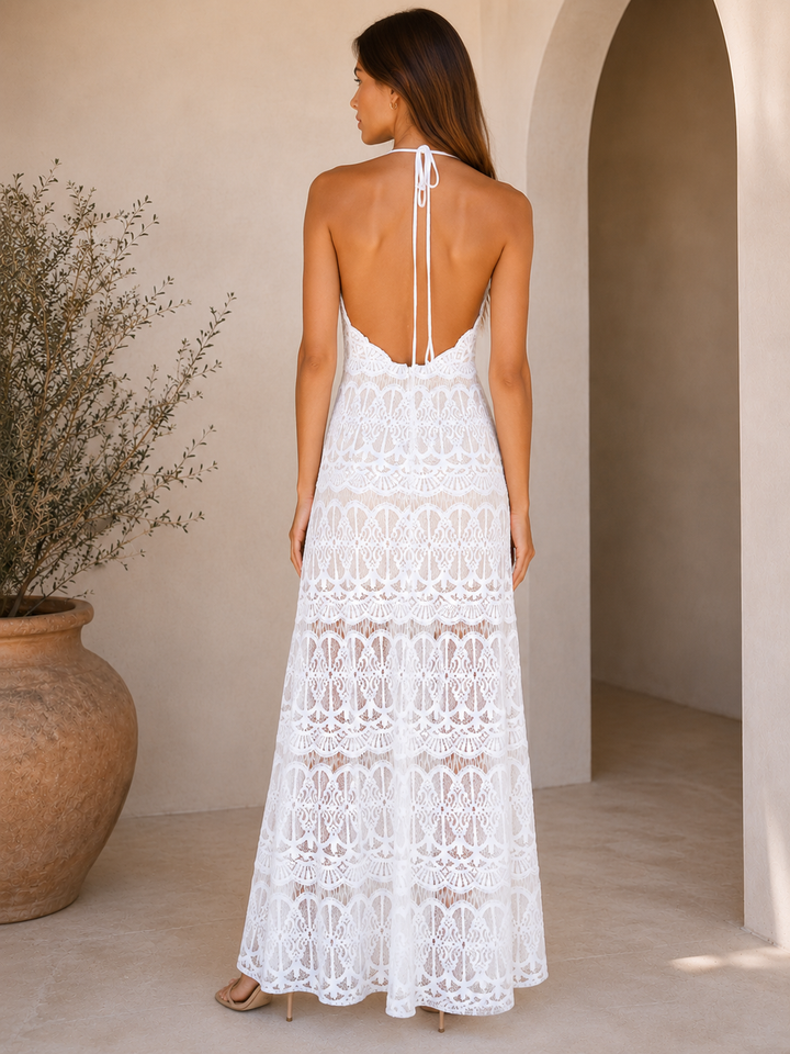 A woman viewed from the back wearing a white lace halter maxi dress with an open back and tie detail, standing in a warm-toned architectural setting with a large terracotta planter nearby.