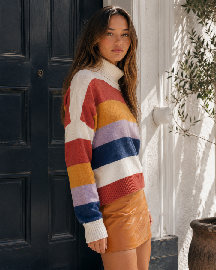 A woman stands in front of a dark paneled door wearing a colorful striped knit sweater with a white turtleneck collar and a glossy tan mini skirt.