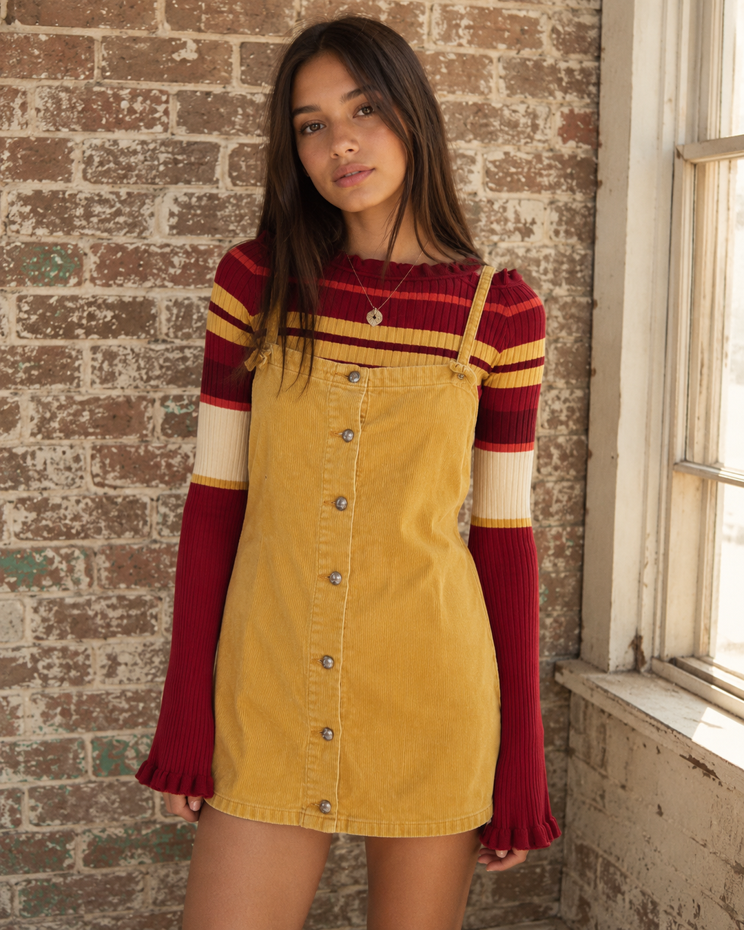Butter yellow corduroy pinafore mini dress with adjustable spaghetti straps and a button-front closure layered over a striped ribbed knit top.

