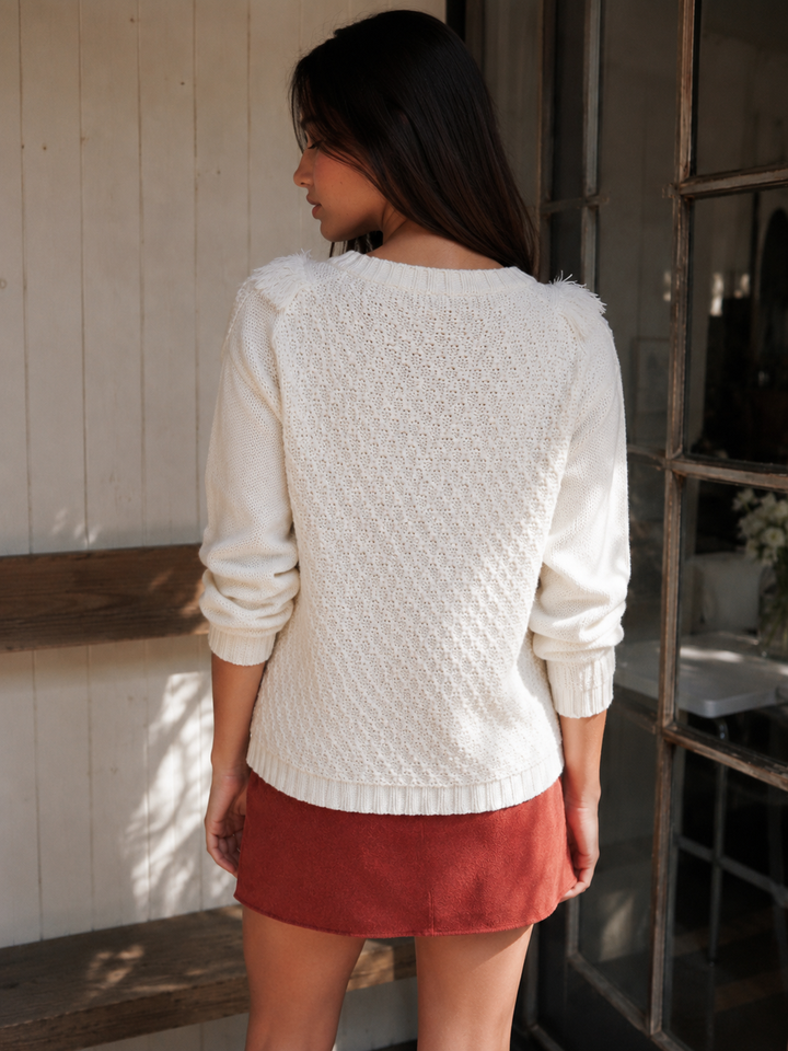 A woman/ wearing a cream textured knit sweater and rust-colored shorts, standing in profile in a rustic room with a white paneled wall and a window.