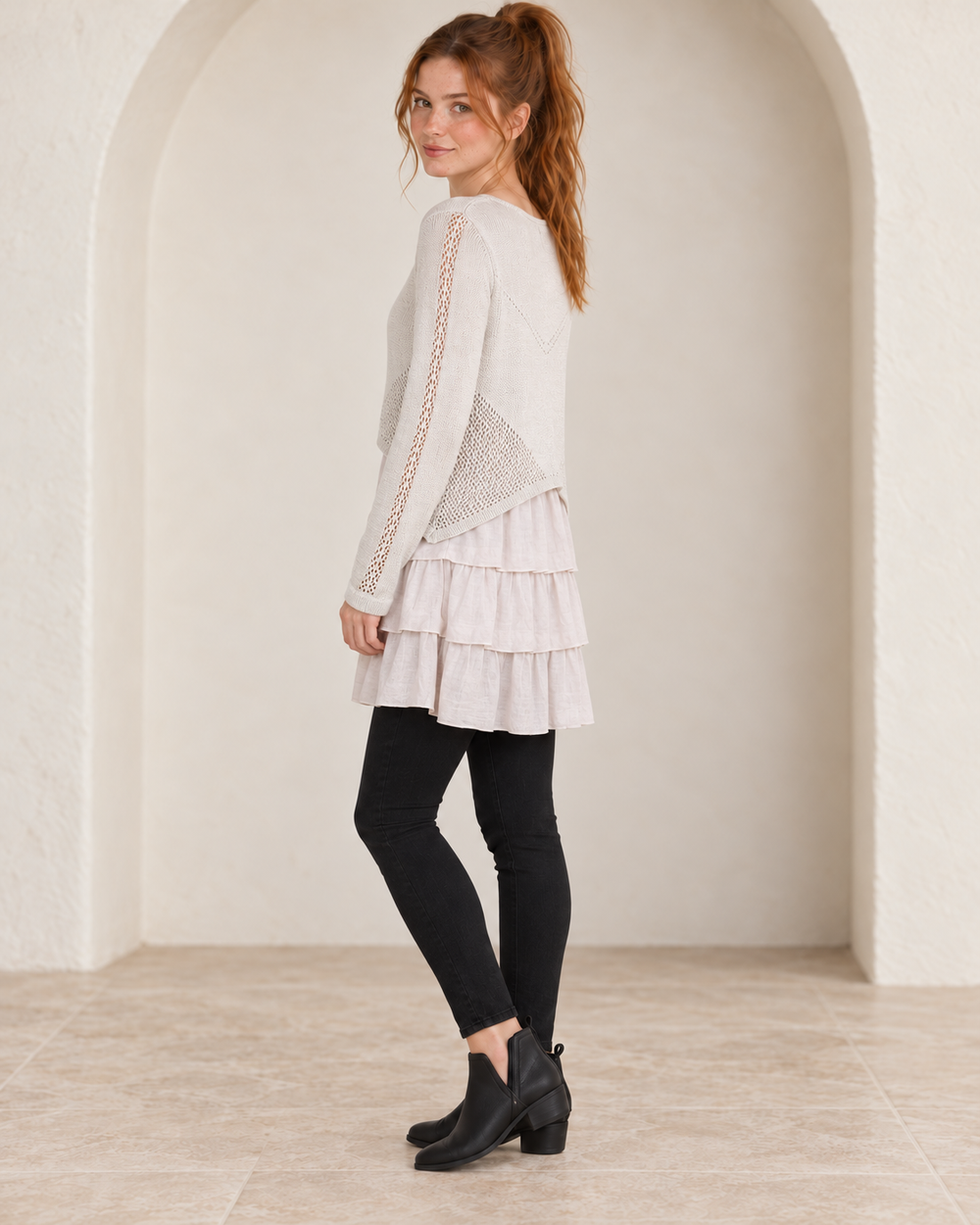 Khaki open-knit ruffle-hem sweater tunic, side-angle view on a model showing openwork sleeves and layered ruffles, styled with black fitted jeans and ankle boots.