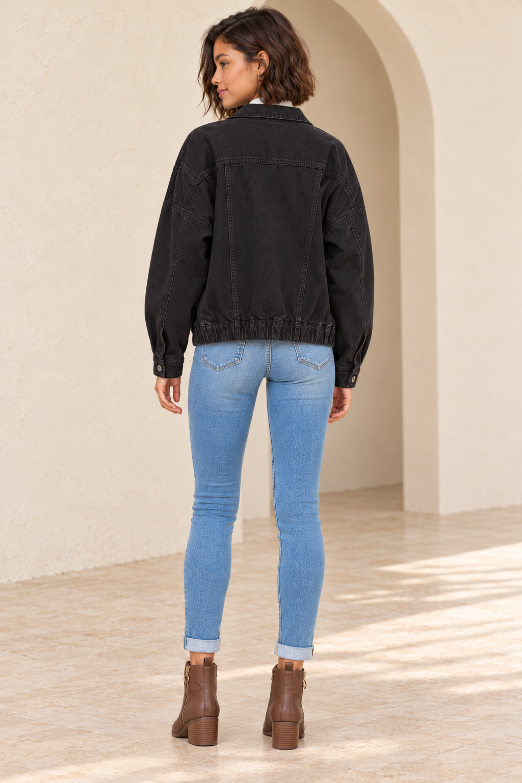 Premium Wash Cotton Denim Jacket in Black, full-length back outfit view showing yoke, two rear panel seams and gathered elastic hem with medium-blue skinny jeans and brown ankle boots.