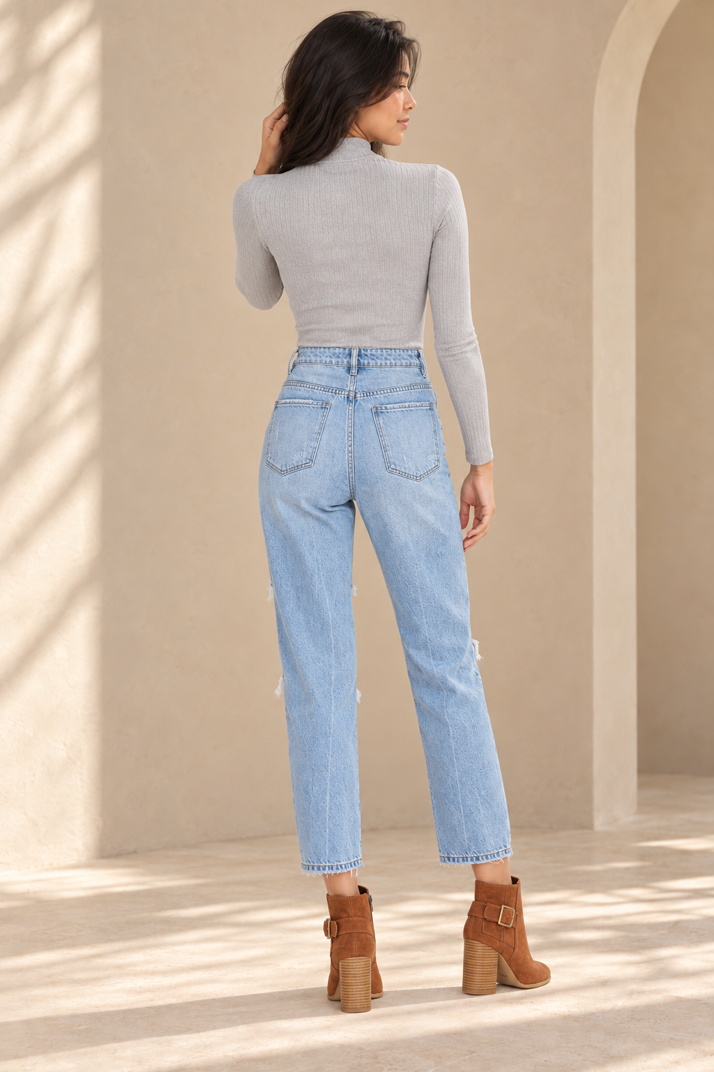 Thalia Distressed High Waisted Denim Mom Jeans in Light Wash, back view on an adult model styled with a heather grey Free People - Lindsey Mock Neck Ribbed Top and cognac Sbicca Lorenza suede ankle booties, in a warm plaster studio.